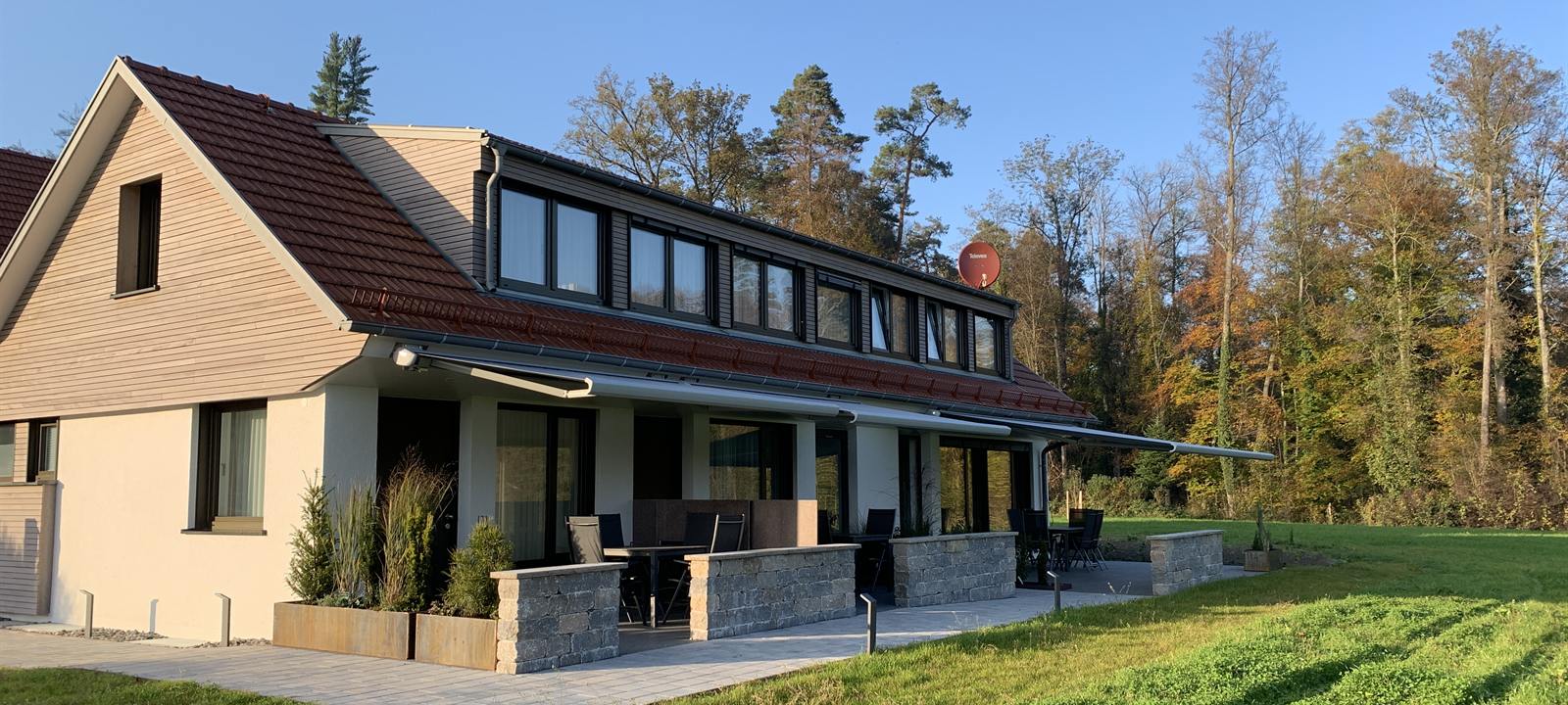 Das Haus im Abendlicht, davor die überdachten Terrassen und die Wiese, dahinter der Wald